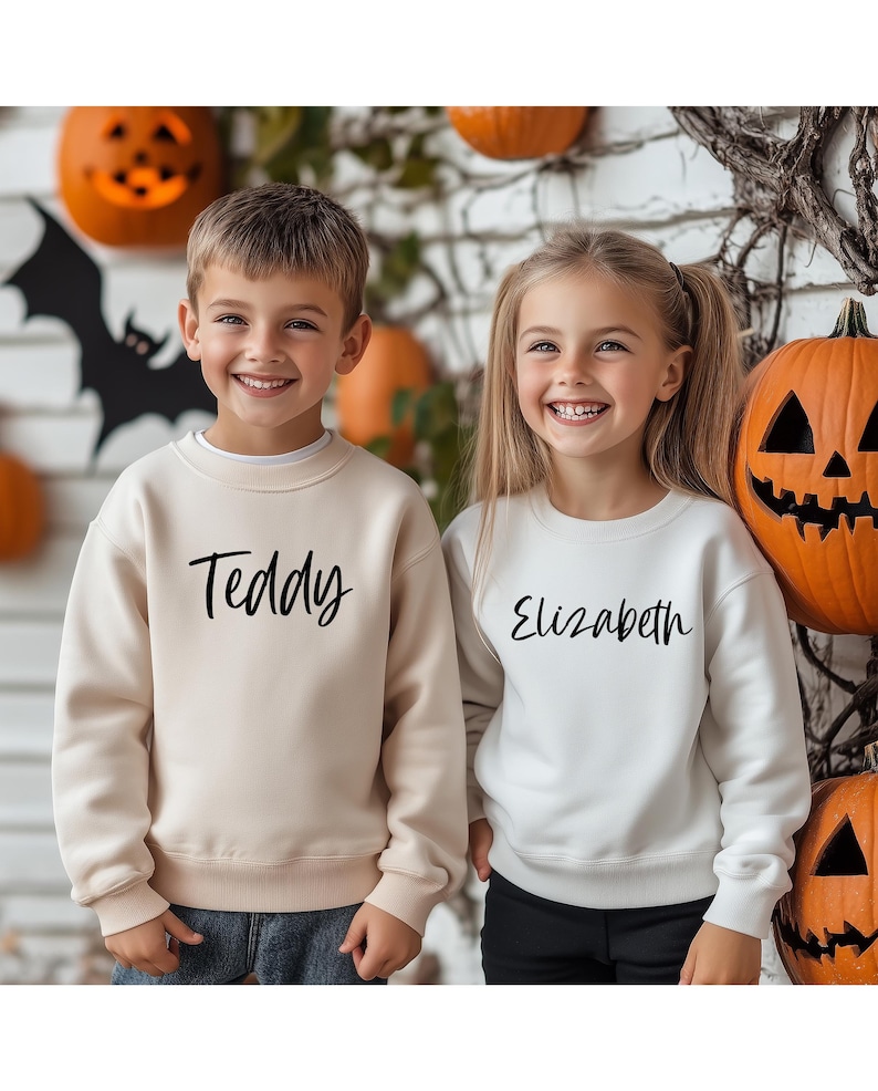 Peut inclure: Deux enfants portant des sweat-shirts cr&egrave;me et blanc avec les noms "Teddy" et "Elizabeth" imprim&eacute;s dessus. Ils sont debout devant un mur de briques avec des citrouilles et des d&eacute;corations d'automne.