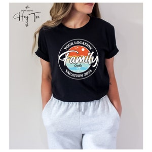 K&ouml;nnte beinhalten: Schwarzes T-Shirt mit einem wei&szlig;en Kreis-Motiv, auf dem "Your Location Family Vacation 2024" steht, wobei das Wort "Enola" im Kreis steht. Das Motiv hat ein Sonnenuntergangsdesign mit Palmen und V&ouml;geln.
