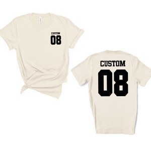 May include: Two cream-coloured t-shirts, one front and one back view. Each shirt has the word "CUSTOM" above the number "08" in black block letters. The front of the shirt has the text on the upper left chest. The back of the shirt has the text centered.