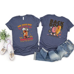 Puede incluir: Dos camisetas azul marino con dise&ntilde;os gr&aacute;ficos. Una camiseta dice "Ask Your Dad About My Throat Game" con un perrito caliente de dibujos animados. La otra dice "Back Door" con un donut y un perrito caliente. Un par de vaqueros y zapatillas blancas tambi&eacute;n est&aacute;n en la imagen.