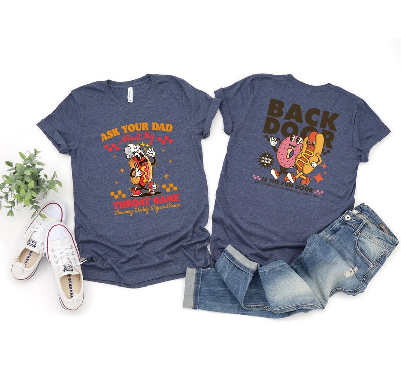 Puede incluir: Dos camisetas azul marino con dise&ntilde;os gr&aacute;ficos. Una camiseta dice "Ask Your Dad About My Throat Game" con un perrito caliente de dibujos animados. La otra dice "Back Door" con un donut y un perrito caliente. Un par de vaqueros y zapatillas blancas tambi&eacute;n est&aacute;n en la imagen.