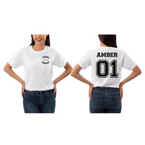 May include: White t-shirts with black text. One shirt reads "TEAM [heart] GRADE" on the front. The other shirt has "AMBER 01" on the back. Both shirts are short-sleeved and worn with blue jeans.