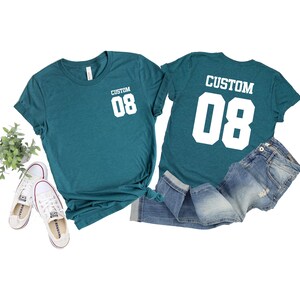 May include: Teal t-shirts with the text "CUSTOM 08" in white, paired with denim jeans and white trainers. One shirt shows the text on the front, the other on the back. A small plant is visible.