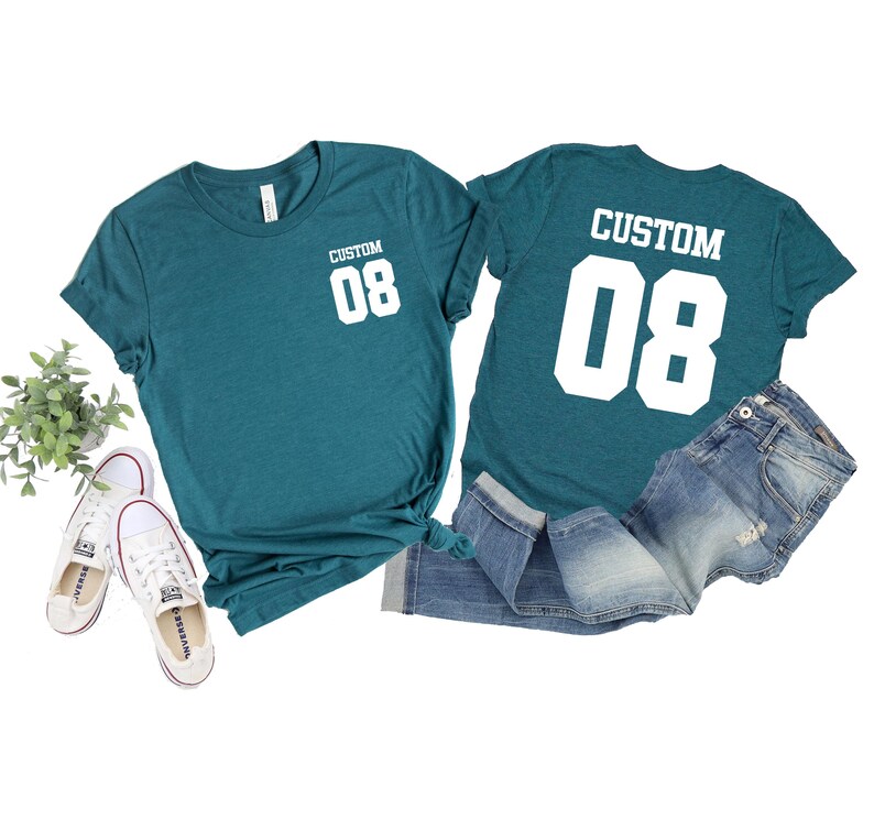 May include: Teal t-shirts with the text "CUSTOM 08" in white, paired with denim jeans and white trainers. One shirt shows the text on the front, the other on the back. A small plant is visible.