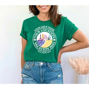 May include: Green t-shirt with a white circle graphic that says "Your Grup Name Your Location Vacation 2023 Sea Sand Sun". The graphic features a beach scene with colorful buildings and a boat.