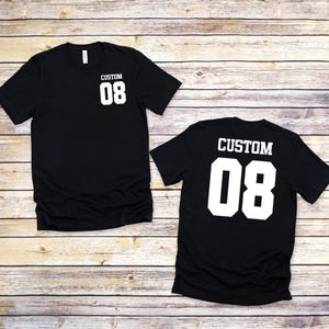 May include: Two black t-shirts with the text "CUSTOM 08" printed in white. One shirt displays the text on the front, the other on the back. The shirts are displayed on a light brown wooden surface.