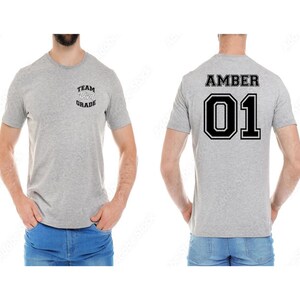 May include: A light gray t-shirt with the text "TEAM Third GRADE" on the front and "AMBER 01" on the back. The shirt is short-sleeved and made of a soft material.