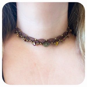 May include: A brown cord choker necklace with a woven design and small gold and burgundy beads. The necklace has a large, round, gold-toned pendant in the center.
