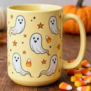 Può includere: Una tazza in ceramica gialla a tema Halloween. La tazza presenta illustrazioni di fantasmi bianchi, stelle arancioni e caramelle a forma di mais. La tazza ha un manico grande ed è circondata da caramelle e una zucca.