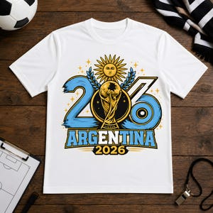 Może przedstawiać: Biały t-shirt z krótkim rękawem z grafiką przedstawiającą liczbę 2026 w kolorze niebieskim, złote trofeum piłkarskie, symbol słońca oraz napis ARGENTINA 2026. Wzór zawiera gałązki laurowe i małe gwiazdki umieszczone na środku przodu.