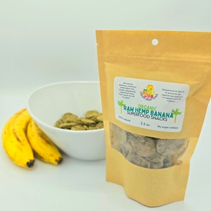 May include: A brown paper bag with a white label that reads "ORGANIC RAW HEMP BANANA SUPERFOOD SNACKS 100% natural 3.5 oz No sugar added". The bag contains dehydrated banana chips. A white bowl with green banana chips is in the background.