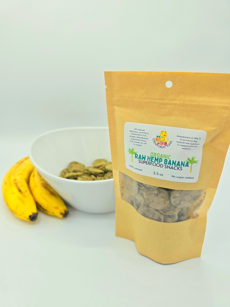 May include: A brown paper bag with a white label that reads "ORGANIC RAW HEMP BANANA SUPERFOOD SNACKS 100% natural 3.5 oz No sugar added". The bag contains dehydrated banana chips. A white bowl with green banana chips is in the background.
