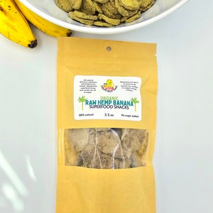 May include: A brown paper bag with a clear window showing a small amount of dried, light brown banana chips. The bag has a label that reads "ORGANIC RAW HEMP BANANA SUPERFOOD SNACKS 100% natural 3.5 oz No sugar added".