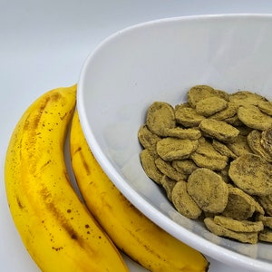 May include: Two yellow bananas next to a white bowl filled with green, round, flat, crunchy snacks.