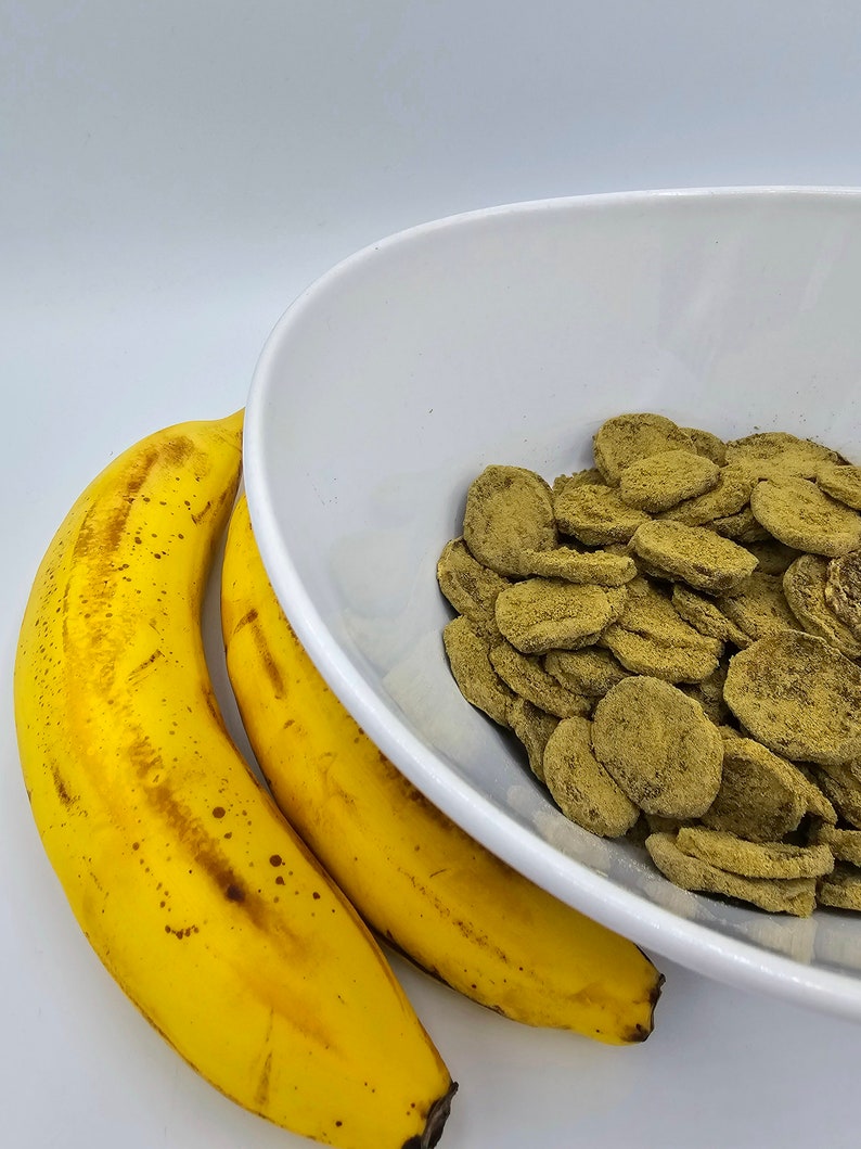 May include: Two yellow bananas next to a white bowl filled with green, round, flat, crunchy snacks.