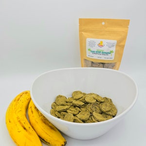May include: A white bowl filled with green hemp and banana superfood snacks. Two yellow bananas are in the foreground. A brown bag with the text "Organic Raw Hemp Banana Superfood Snacks 3.5 oz No sugar added" is in the background.