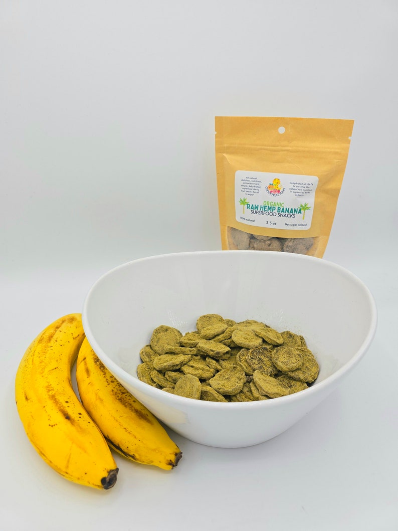 May include: A white bowl filled with green hemp and banana superfood snacks. Two yellow bananas are in the foreground. A brown bag with the text "Organic Raw Hemp Banana Superfood Snacks 3.5 oz No sugar added" is in the background.