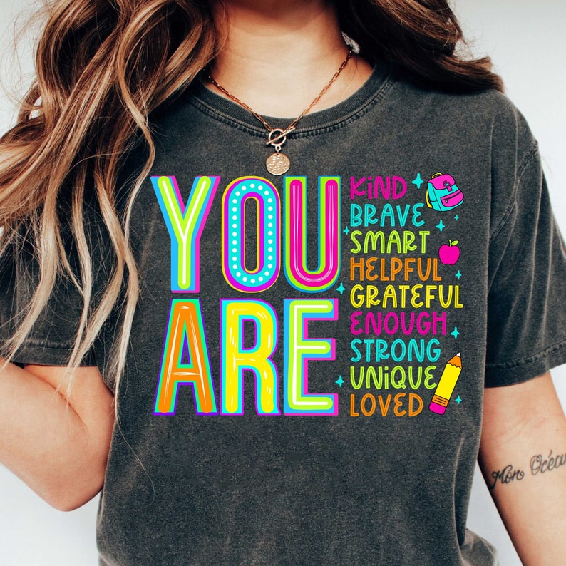 Pu&ograve; includere: Una maglietta grigia scura con le parole "YOU ARE" in grandi lettere colorate. Le parole "KIND, BRAVE, SMART, HELPFUL, GRATEFUL, ENOUGH, STRONG, UNIQUE, LOVED" sono impilate verticalmente in lettere pi&ugrave; piccole attorno al testo principale. Ci sono piccole illustrazioni di uno zaino, una mela e una matita.