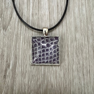 May include: A square pendant necklace with a black cord. The pendant is silver with a clear resin insert that shows a purple snakeskin pattern.