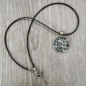 May include: A black leather cord necklace with a round silver pendant featuring a black and white snake skin pattern.