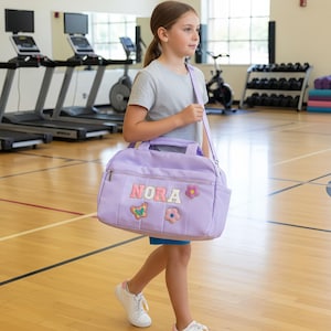 Puede incluir: Una bolsa de deporte lila con el nombre "NORA" en letras coloridas y parches decorativos. La bolsa tiene una correa para el hombro y un asa superior. La bolsa es llevada por una persona en un gimnasio.
