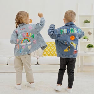May include: Two children's denim jackets, light blue in colour, adorned with colourful patches and names. One jacket features the name "MELIA" with rainbow and unicorn motifs. The other displays "FORREST" with basketball and dinosaur patches.