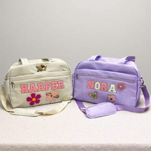 May include: Two personalized diaper bags. One is cream-colored with the name "HARPER" in pink letters, a flower, and a winged unicorn. The other is lavender with "NORA" in pink letters, a butterfly, and a flower.