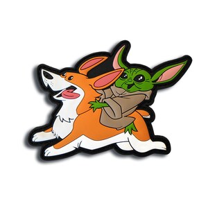 May include: A colorful patch featuring a cartoon corgi dog with an open mouth and a green alien character riding on its back. The patch has a black border and is set against a white background.
