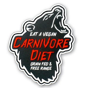May include: A black patch shaped like a roaring bear head with white outlines. The text reads EAT A VEGAN in white, CARNIVORE DIET in red, and GRAIN FED & FREE RANGE in white. The design features a textured black surface with bold, raised lettering.