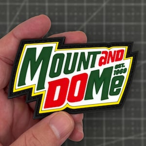 May include: A rectangular rubber patch with a black border and yellow outline. The text reads MOUNT AND DOME in bold green and red letters, with EST. 1969 in small green text on the right side. The design features a jagged, multi-layered shape.