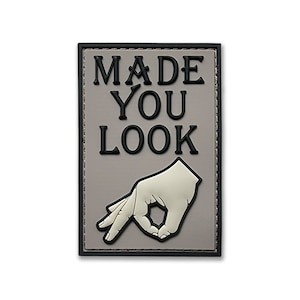 May include: Rectangular patch with the words "MADE YOU LOOK" in black, raised lettering. Below the text is a white hand gesture, also raised. The patch has a gray background and a black border.