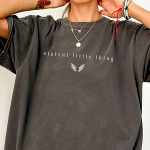 May include: Dark gray t-shirt with the text "violent little thing" and a bat wing graphic. The shirt has a vintage, worn look and a relaxed fit. The text and graphic are in a muted, light color.