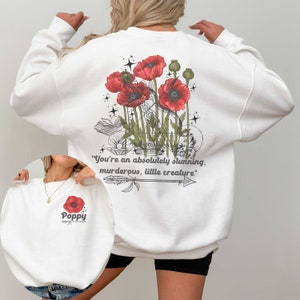 Pode incluir: Camisola branca com um gráfico de flor de papoula vermelha e o texto "You're an absolutely stunning, murderous, little creature." nas costas. A frente da camisola tem um gráfico de flor de papoula menor e a palavra "Poppy" por baixo.
