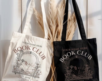 Może przedstawiać: Dwie torby, jedna kremowa i jedna czarna, z napisem "YOUR NAME HERE BOOK CLUB EST. 2024". Torby mają kwiatowy wzór wokół łukowatej ramy. Torby mają długie uchwyty.