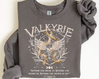 Może przedstawiać: Szara bluza z nadrukiem graficznym przedstawiającym czaszkę z skrzydłami i słowem "VALKYRIE" w stylu vintage. Grafika otoczona jest wieńcem z kwiatów i liści. Tekst pod grafiką brzmi "NOTHING CAN BREAK US. NOTHING CAN SILENCE US. NOTHING CAN DROWN US OUT."