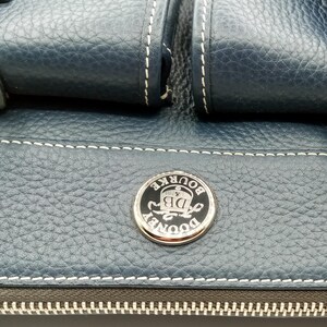 May include: Close-up of a navy blue leather handbag with white stitching. The bag features a silver and black circular emblem with the text "DOONEY & BOURKE DB". The zipper is visible at the bottom.
