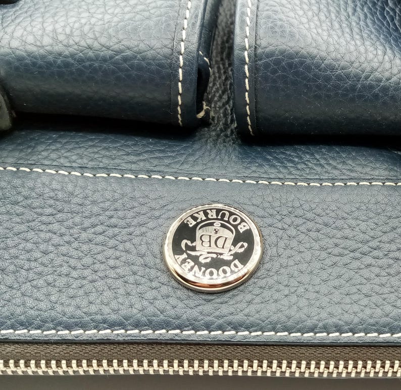 May include: Close-up of a navy blue leather handbag with white stitching. The bag features a silver and black circular emblem with the text "DOONEY & BOURKE DB". The zipper is visible at the bottom.