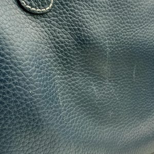 May include: Close-up of a dark blue leather bag. The leather has a textured, pebbled appearance with visible grain and subtle variations in color. White stitching is visible on the handle attachment. The bag's surface shows some wear.