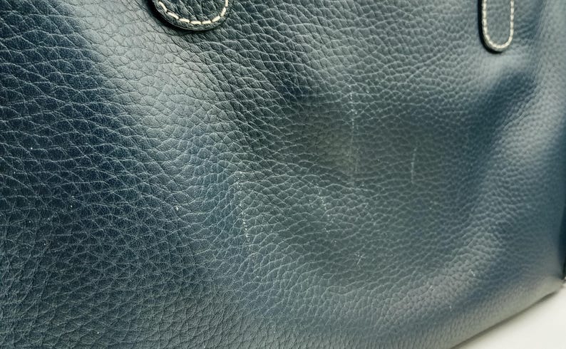 May include: Close-up of a dark blue leather bag. The leather has a textured, pebbled appearance with visible grain and subtle variations in color. White stitching is visible on the handle attachment. The bag's surface shows some wear.