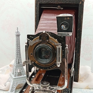 May include: Vintage Eastman Kodak Company camera with a brown leather bellows and a black lens. The camera has a silver metal frame and a small viewfinder. The camera is open, revealing the lens and internal components. A silver Eiffel Tower model is to the left.