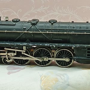 May include: A black metal toy steam locomotive train engine featuring a detailed boiler, a cab with windows, and silver-toned metal wheels connected by rods. The paint shows signs of wear, revealing the metallic texture of the model train.