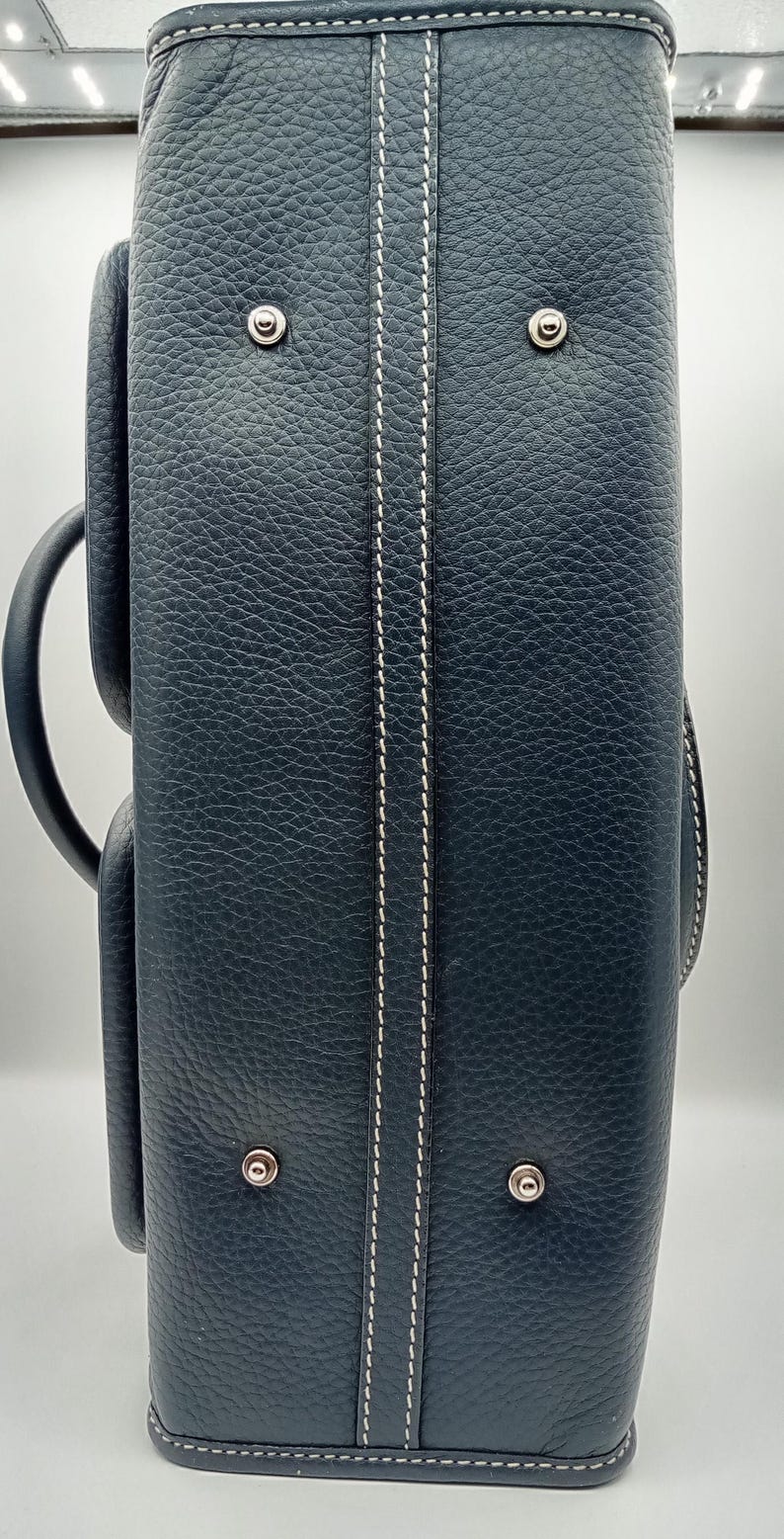May include: A navy blue leather briefcase with white stitching details. The briefcase has a textured surface, a handle, and metal hardware. The design includes a vertical white stitch detail down the center.