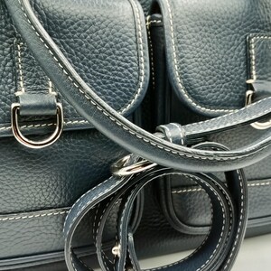 May include: Dark blue leather handbag with white stitching and silver-tone hardware. Features two front pockets, a top handle, and a detachable shoulder strap. The bag has a textured finish and a metal logo emblem.