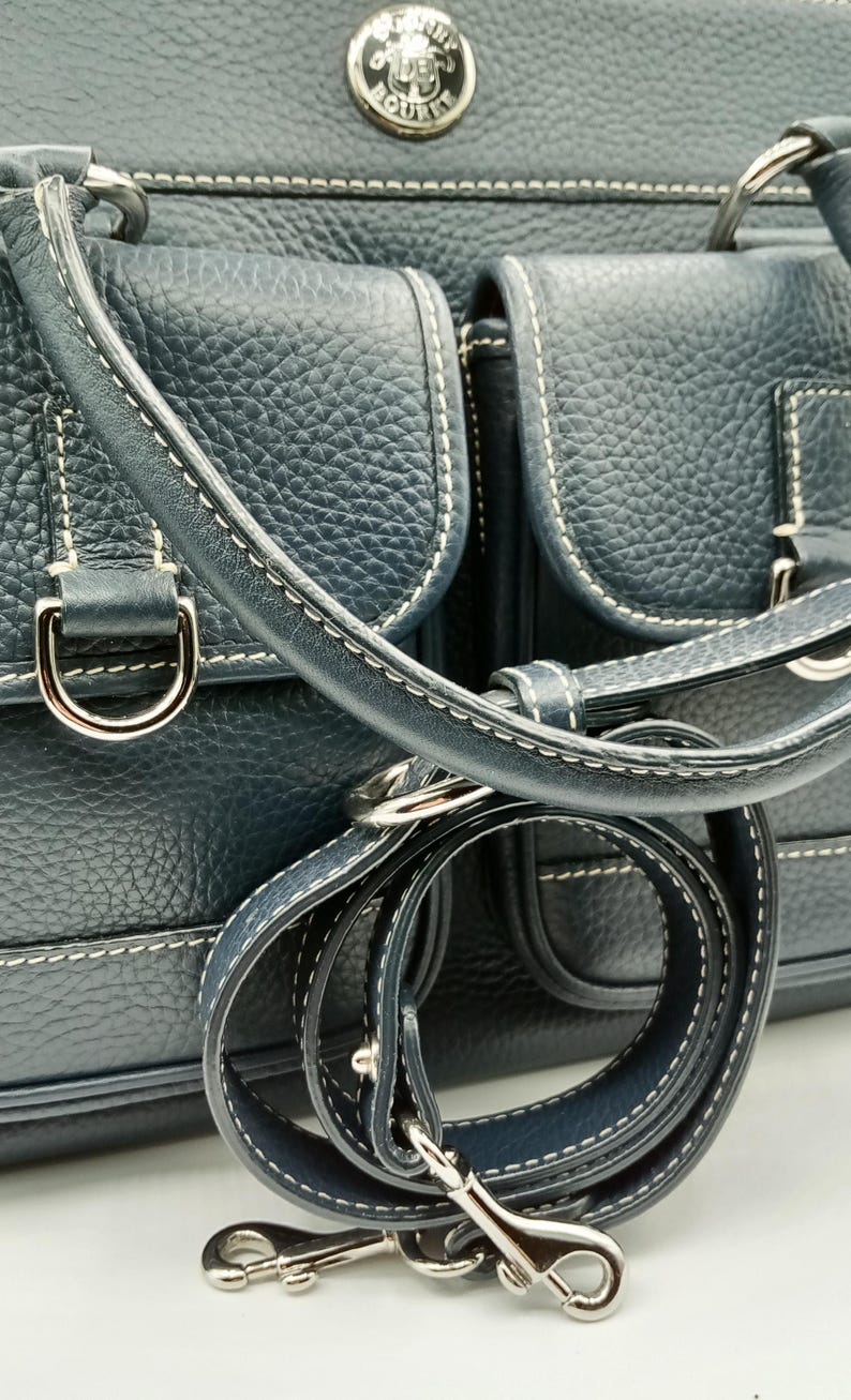 May include: Dark blue leather handbag with white stitching and silver-tone hardware. Features two front pockets, a top handle, and a detachable shoulder strap. The bag has a textured finish and a metal logo emblem.