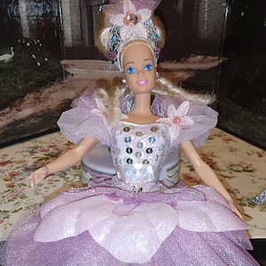 May include: A doll with blonde hair styled in an updo, wearing a silver and pink crown with floral accents. The doll is dressed in a lavender gown featuring a bodice with silver sequins, sheer puff sleeves, and a layered skirt with petal-shaped fabric.