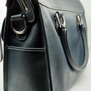 May include: A navy blue leather handbag with a textured finish. The bag features a silver zipper, white stitching, and silver hardware on the handles. The bag has a structured shape and a pocket on the side.