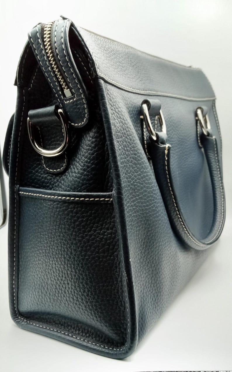 May include: A navy blue leather handbag with a textured finish. The bag features a silver zipper, white stitching, and silver hardware on the handles. The bag has a structured shape and a pocket on the side.