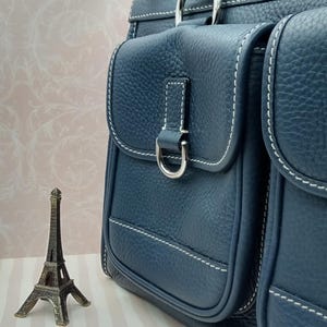 May include: A navy blue leather handbag with two front pockets, white stitching, and silver hardware. The bag has a top handle and a decorative metal emblem. A small Eiffel Tower figurine is in the foreground.