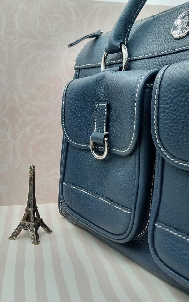 May include: A navy blue leather handbag with two front pockets, white stitching, and silver hardware. The bag has a top handle and a decorative metal emblem. A small Eiffel Tower figurine is in the foreground.