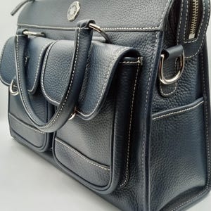 May include: A navy blue leather satchel with white stitching and multiple exterior pockets. The bag features a top zipper closure, silver-tone hardware, and a handle for carrying. A small emblem is visible on the front.